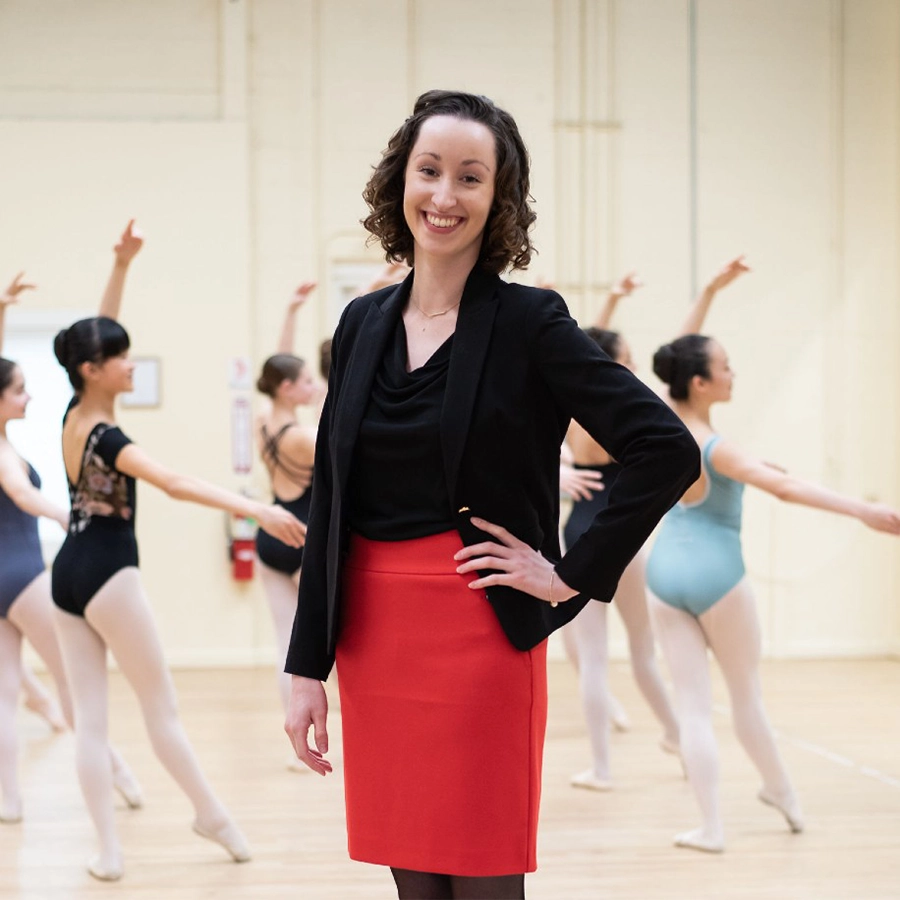A woman poses, hands on hip, while smiling at the camera inside a dance studio filled with ballerinas.